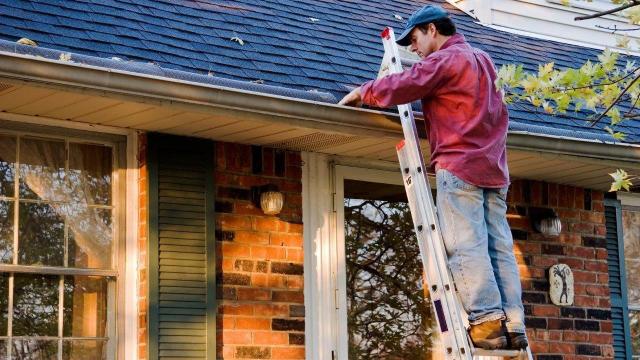 A person is on a ladder inspecting or repairing the gutter of a brick house with a shingle roof.