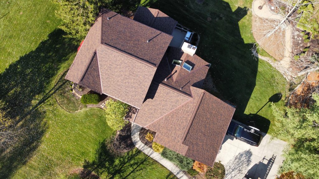 An aerial view of a large house by a lumbus Ohio Roofing Contractor with a brown roof surrounded by trees and a lawn with a driveway on the side.