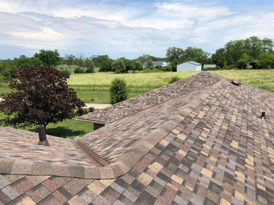 The image shows a patterned shingle rooftop with a scenic view of green fields and trees in the background.