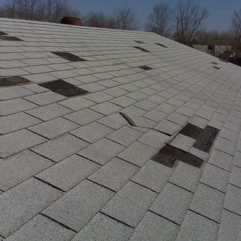 A damaged shingle roof with multiple missing pieces exposing the underlayment, in need of repair.