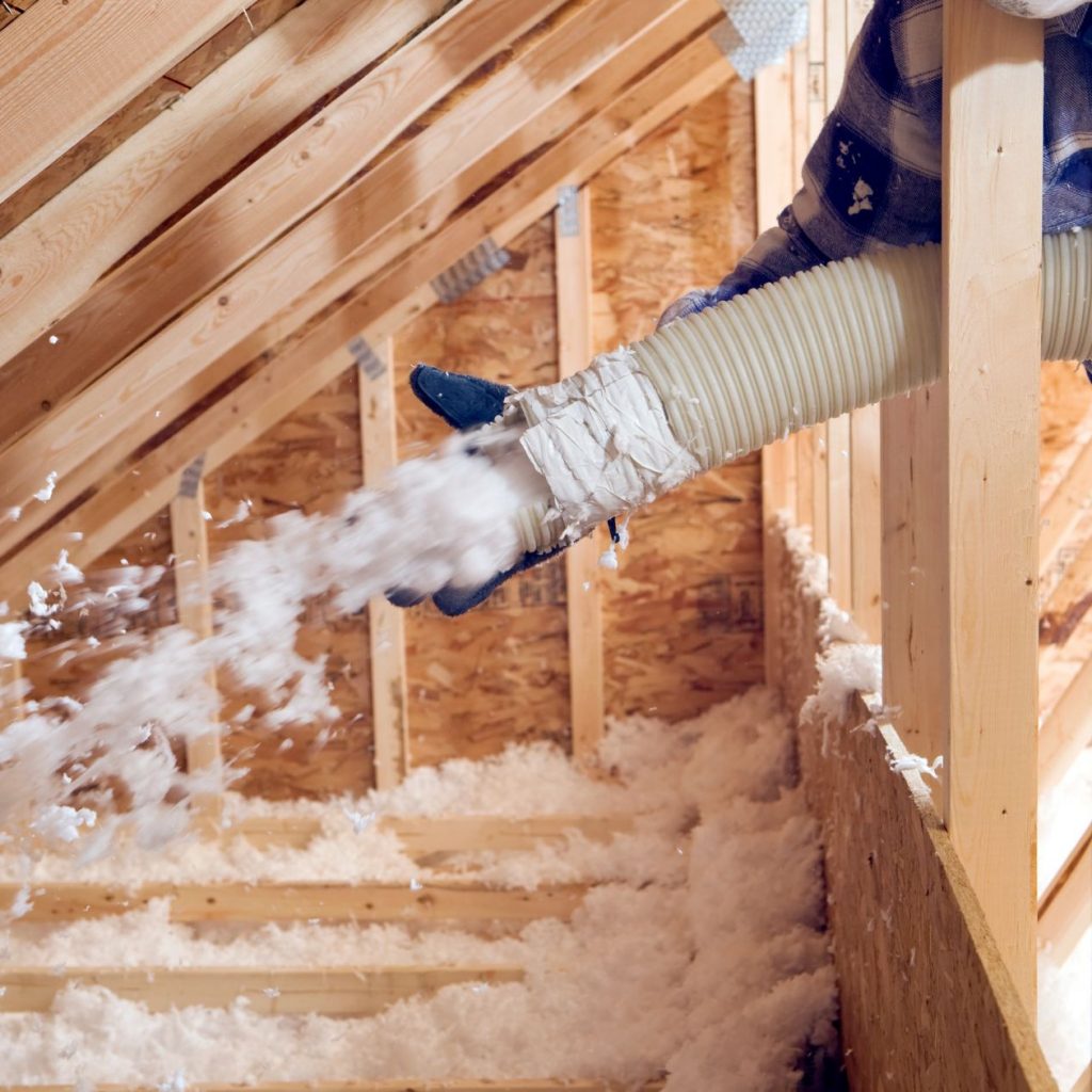A worker is installing blown-in insulation in the attic of a house between wooden rafters.