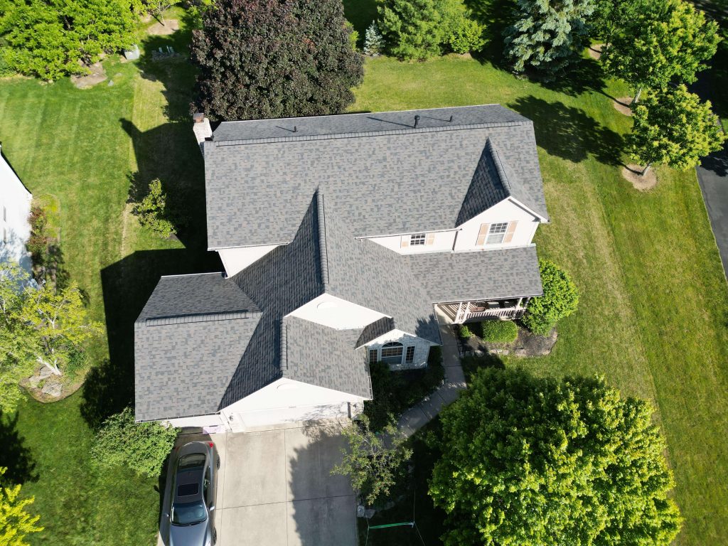 Aerial view of a two-story house with a gray roof, surrounded by greenery, with a car parked in the driveway. Upper arlington  OH roofing contractor
